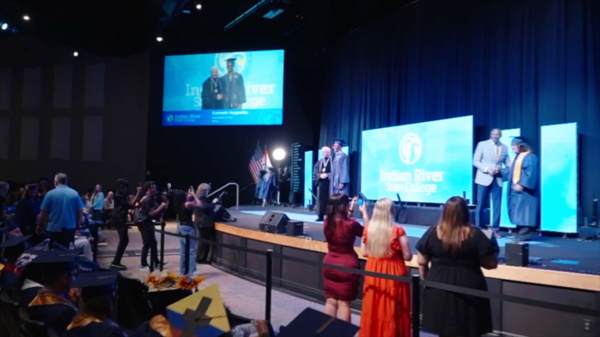 A graduate being honored on stage in front of an IRSC-branded LED screen